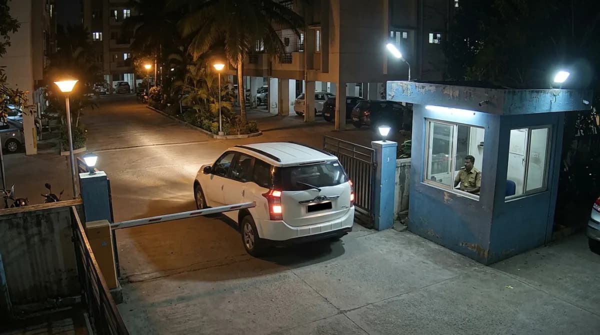 Night-time security camera view of a white SUV stopped at an apartment gate barrier