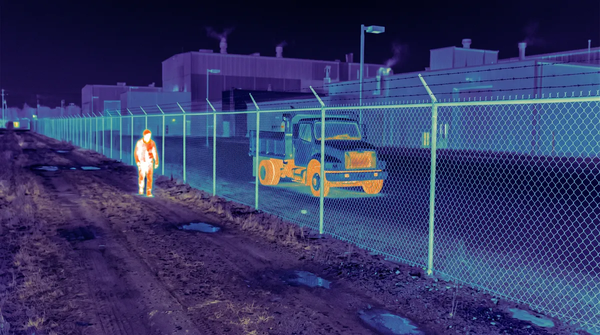 Thermal camera view of a person as a bright heat silhouette beside a factory fence at night